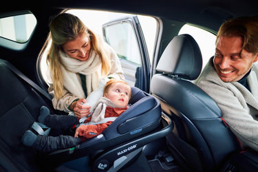 Mother placing baby in a Maxi-Cosi in the car, father sitting in the front seat